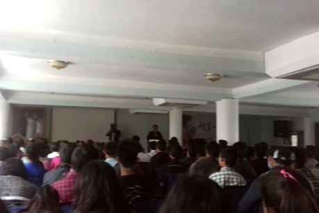 A full hall seen from the back, the audience facing a speaker at the front